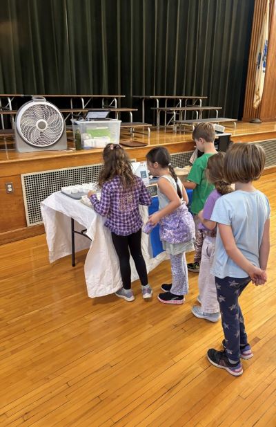 A group of children line up at a covered table inside a gymnasium with wooden floors.